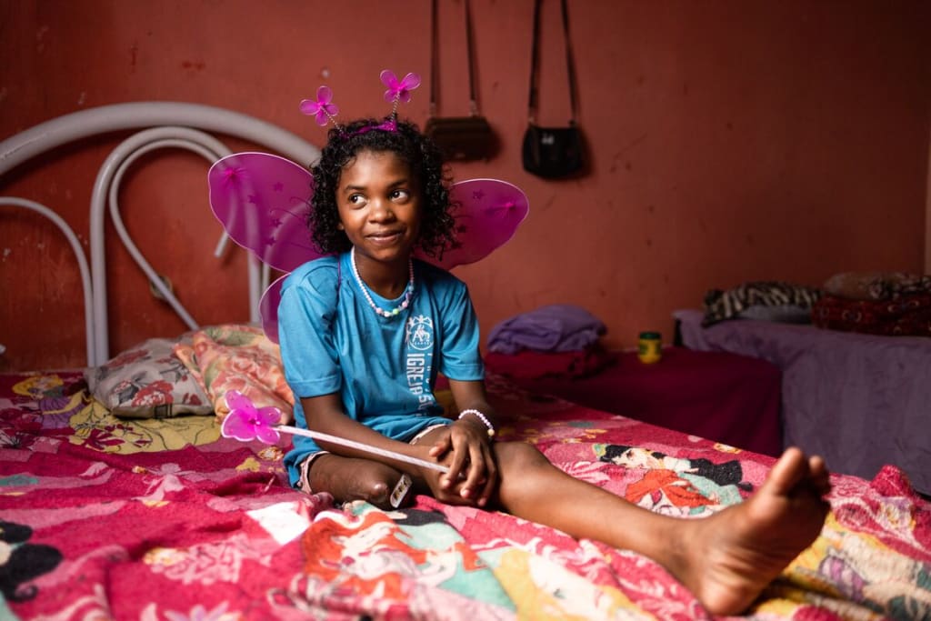 A girl in a blue shirt and pink wings sits on a bed in a pink room